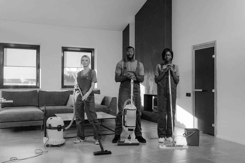 Three professional cleaners from Queens Park Carpet Cleaners are engaged in a surface cleaning process inside a modern living room, which features a large L-shaped sofa, two windows letting in natural