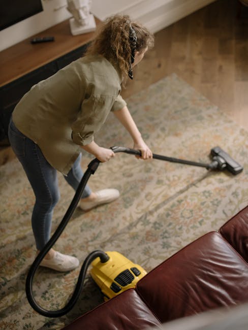 A yellow handheld vacuum cleaner with a flexible hose and a metal extension wand is positioned on an ornate patterned rug in a living room. The room features wooden flooring, a black metal and wooden